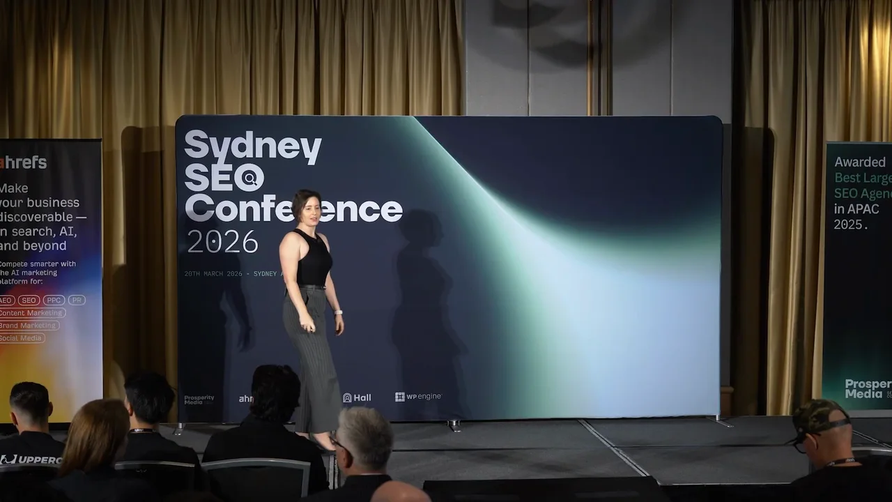 Jes Scholz speaking on stage at Sydney SEO Conference 2026 in front of the event backdrop