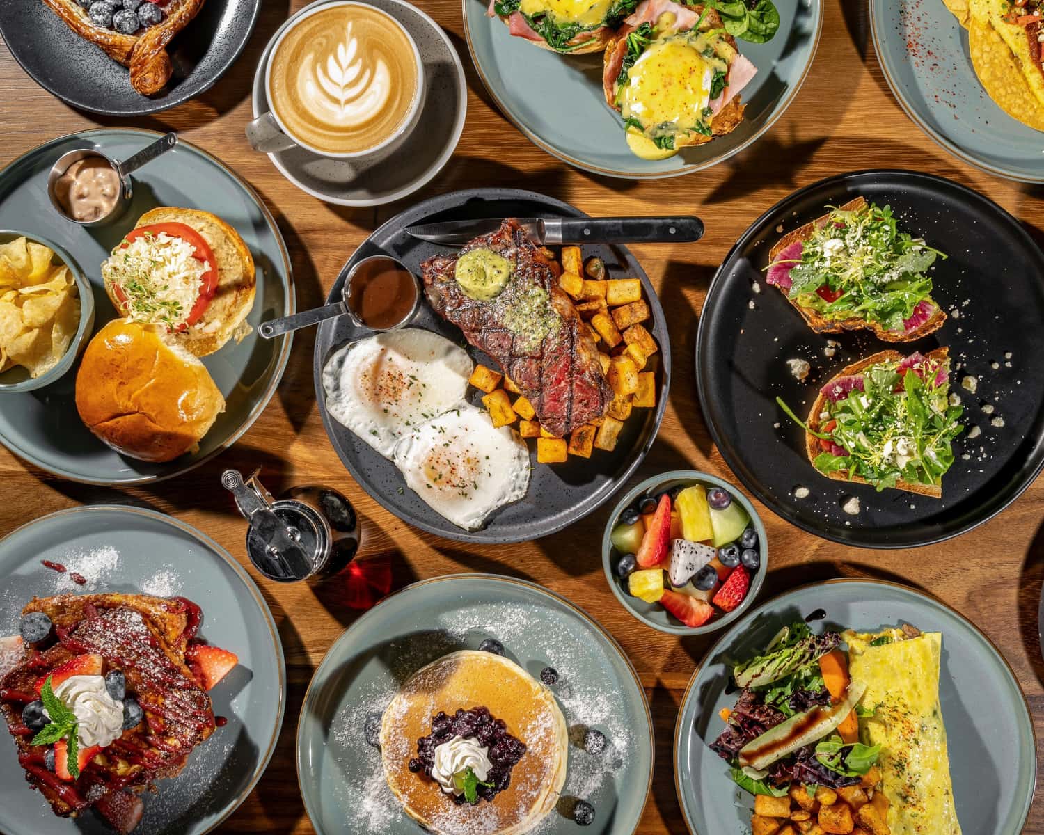 Assortment of breakfast dishes including steak, eggs, pancakes, sandwiches, and coffee on wooden table for Kensington spots